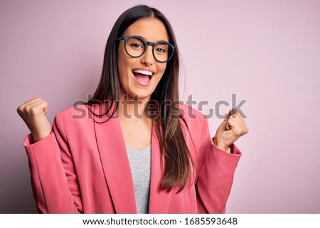 Young beautiful brunette businesswoman wearing jacket and glasses over pink background celebrating surprised and amazed for success with arms raised and open eyes. Winner concept.