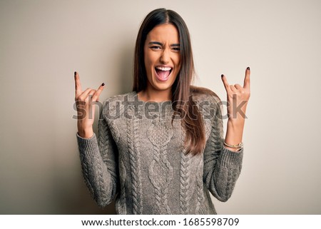 Young beautiful brunette woman wearing casual sweater over isolated white background shouting with crazy expression doing rock symbol with hands up. Music star. Heavy concept.