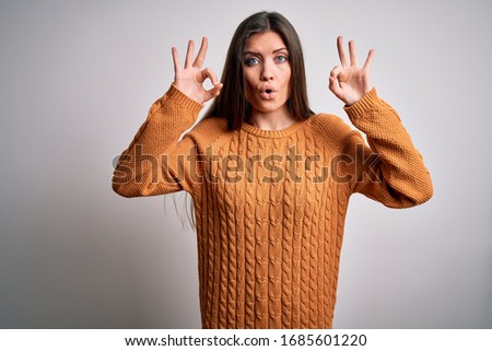 Young beautiful woman with blue eyes wearing casual sweater standing over white background looking surprised and shocked doing ok approval symbol with fingers. Crazy expression
