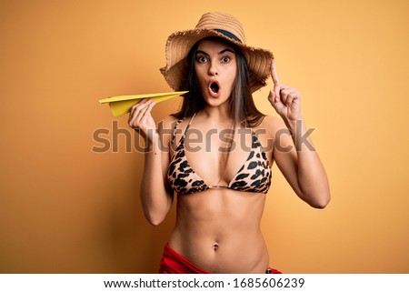 Young beautiful woman on vacation wearing bikini and summer hat holding paper airplane surprised with an idea or question pointing finger with happy face, number one