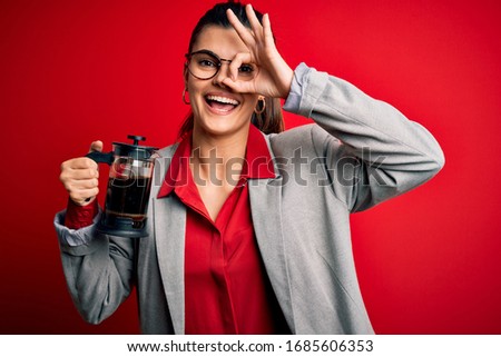 Young beautiful brunette woman doing coffe holding french coffeemaker over red background with happy face smiling doing ok sign with hand on eye looking through fingers