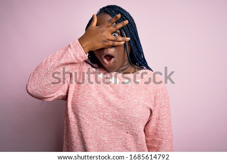 African american plus size woman with braids wearing casual sweater over pink background peeking in shock covering face and eyes with hand, looking through fingers with embarrassed expression.
