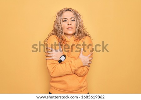 Young beautiful blonde sporty woman wearing casual sweatshirt over yellow background shaking and freezing for winter cold with sad and shock expression on face