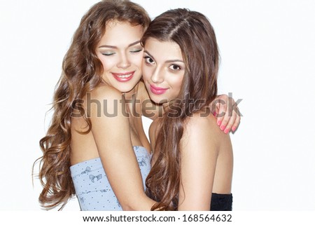 Pretty brunette girls with bright makeup and pink lips having fun. Girl with closed eyes hugging her friend. Other smiling and looking at camera. Inside. Wearing dresses.