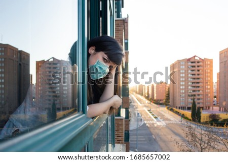 Bored woman with ffp2 face mask peeks out the window during quarantine over covid-19 crisis. Stay at home concept. Empty city under confinement. Valladolid, Spain. Royalty-Free Stock Photo #1685657002