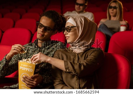 African american muslim man with his wife sitting in movie theater, watching 3D movie, eating popcorn, smiling.