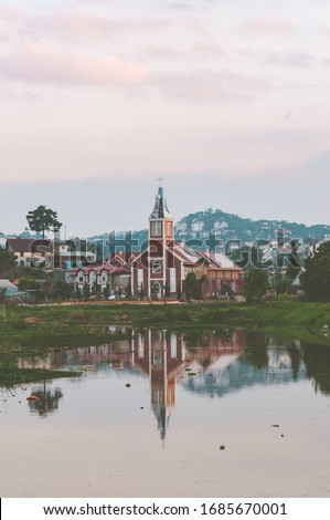 Outdoor landscape photo of a small church with a broken bell tower on the bank of a small lake. Reflection of the colorful twilight sky and the church building create a beautiful scene. Dalat, Vietnam