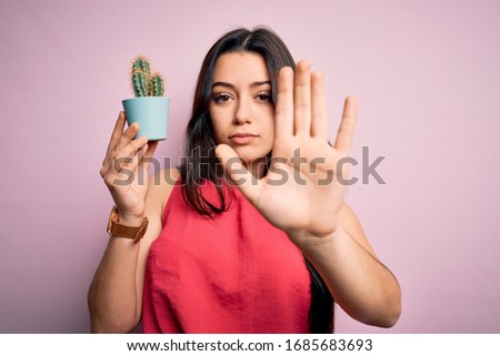 Young brunette woman holding succulent cactus plant over pink background with open hand doing stop sign with serious and confident expression, defense gesture