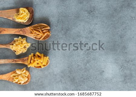 Different types of pasta lying in wooden spoons lying on grey table. Penne, tortellini, fuzilli, and farfalle. Copy space. Horizontal. Royalty-Free Stock Photo #1685687752