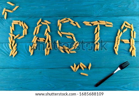 Word pasta laid out of raw pasta penne with fork on blue wooden background