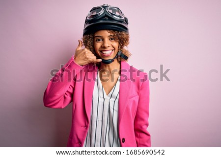 African american motorcyclist woman with curly hair wearing moto helmet over pink background smiling doing phone gesture with hand and fingers like talking on the telephone. Communicating concepts.