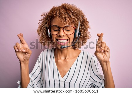 African american curly call center agent woman working using headset over pink background gesturing finger crossed smiling with hope and eyes closed. Luck and superstitious concept.