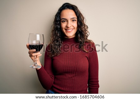 Young beautiful woman with curly hair drinking glass of red wine over white background with a happy face standing and smiling with a confident smile showing teeth