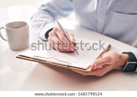 Close-up of unrecognizable businessman sitting at table with tea mug and making notes in application form Royalty-Free Stock Photo #1685798524