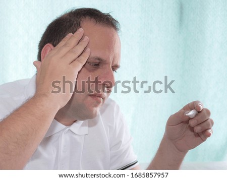 detail of middle aged man touching his head and looking at the thermometer because he is ill