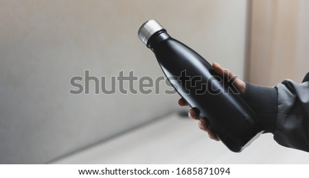 Close-up of female hand holding reusable eco steel thermo water bottle of black on background of office table. Royalty-Free Stock Photo #1685871094