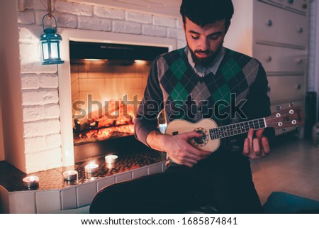 Picture of young man sitting at fireplace and playing ukulele. Alone in room inside practicing. Singing songs and playing melodies.