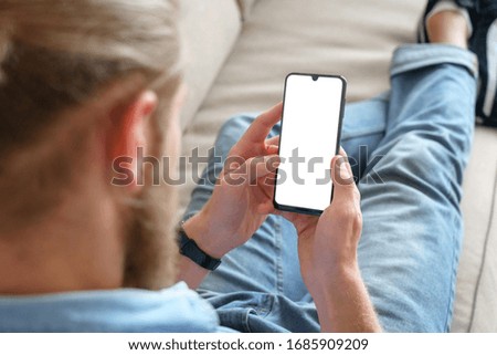 Young male tech user relaxing on sofa holding smartphone mock up blank white screen. Man customer using cell phone mobile social media or shopping app, reading news, sport betting. Over shoulder view