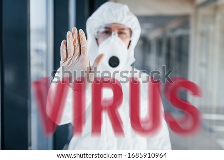 Red virus word. Nurse in protective uniform, mask and glasses standing indoors.