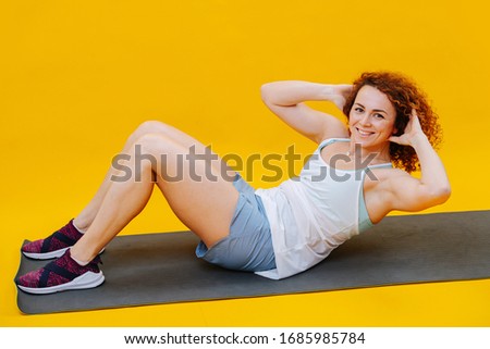 The girl lying on the mat does exercises on the abdominal muscles. She's in sportswear, pictured on a yellow background