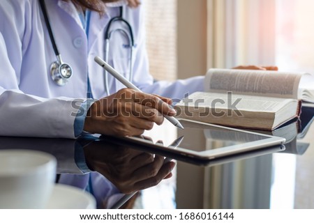 Female doctor in white lab coat reading book and work on digital tablet  computer on the desk at office in medical room at clinic or hospital. Online medical learning concept. Royalty-Free Stock Photo #1686016144