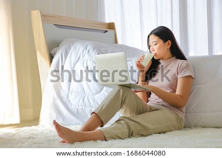 Young pretty girl working from home on her bed using computer laptop to access her work online while drinking a glass of milk during the time of social distancing policy with copy space