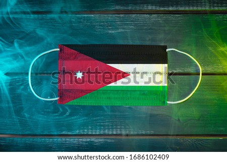 Medical mask for protection against airborne diseases, painted in the national flag of Jordan on a black background in blue-green smoke. Medical protection against airborne diseases, coronavirus