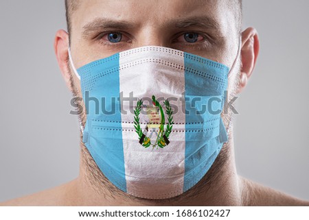 Young man with sore eyes in a medical mask painted in the colors of the national flag of Guatemala. Medical protection against airborne diseases, coronavirus. Man is afraid of getting the flu