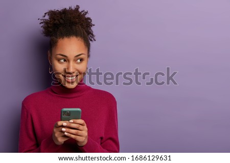 Smiling pleasant looking woman receives message from dating app, holds mobile phone in hands, looks happily aside, edits photo to post online, has curly hair, isolated on violet wall, copy space