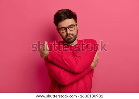 Gorgeous calm bearded man hugs himself, keeps arms around his shoulders, keeps eyes closed, wears soft red jumper, transparent glasses, isolated on pink background, feels coziness, being chilly
