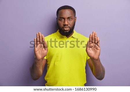 Stop right here. Serious African American man pulls hand towards camera in prohibition gesture, forbids something, dressed in casual clothes, poses against purple background, doesnt permit you