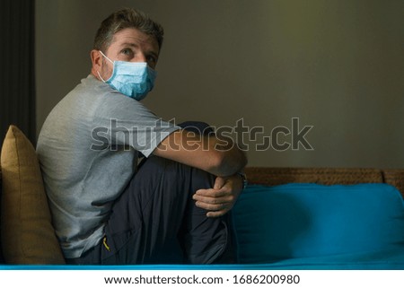 covid-19 virus lockdown - sad and worried man on his 30s or 40s covered with medical mask thinking and feeling scared in quarantine following stay at home instructions to contain virus pandemic