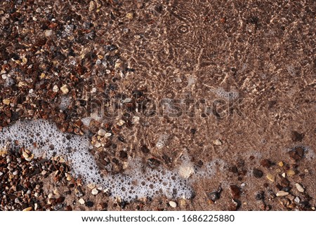 Close-up picture of sandy beach sand with small colorful stones rocks and foamy waves of sea water. Natural texture tropical background.