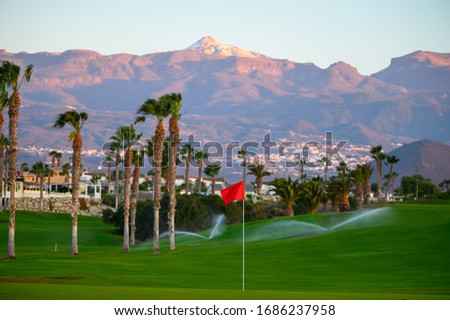 View on evergreen grass field with red pin on large golf course and mountains on tropical island on sunrise