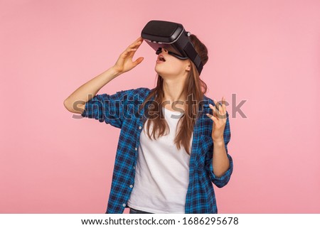 Portrait of gamer girl in checkered shirt wearing vr headset, playing virtual reality game with amazed facial expression, surprised by innovative technology. studio shot isolated on pink background