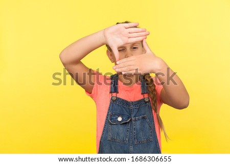 Portrait of little girl in denim overalls looking through photo frame hand gesture, focusing zooming picture, child observing world with interest. indoor studio shot isolated on yellow background