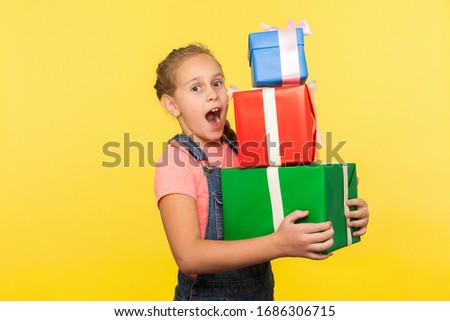 Portrait of surprised little girl with braid in denim overalls holding lot of gift boxes and looking with amazement, shocked by many presents on her birthday. studio shot isolated on yellow background