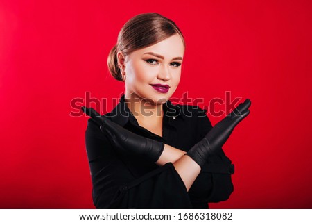 Beautiful girl beautician with collected hair shows a no gesture. Hands in disposable black gloves. Woman on a red background. Studio photography
