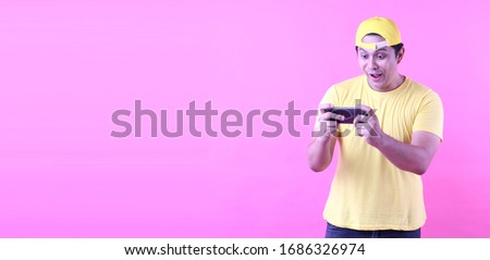 Asian Man wearing a yellow hat .fun addicted to play mobile game.on pink background in studio With copy space