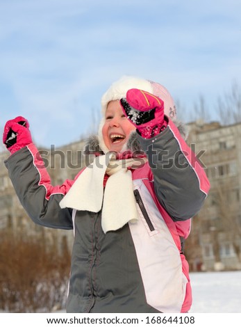 Beautiful vivacious laughing little girl cheering with her hands raised as she plays in the fresh snow on a cold winter day