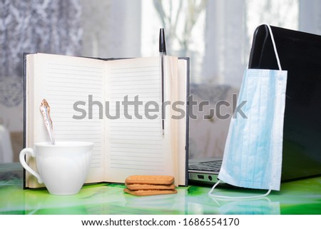 Notebook with a ballpoint pen, a laptop, a Cup of coffee or tea, a cookie stands on the table at home with a protective mask on the background of a soft sofa and a window, education, work, business, s