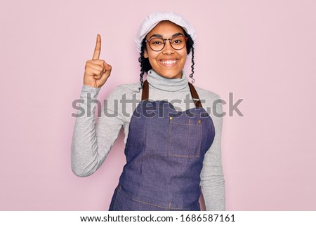 Young african american curly baker woman wearing apron and cap over pink background pointing finger up with successful idea. Exited and happy. Number one.