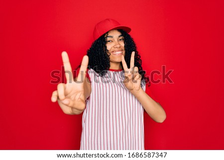 Young african american curly sportswoman wearing baseball cap and striped t-shirt smiling looking to the camera showing fingers doing victory sign. Number two.