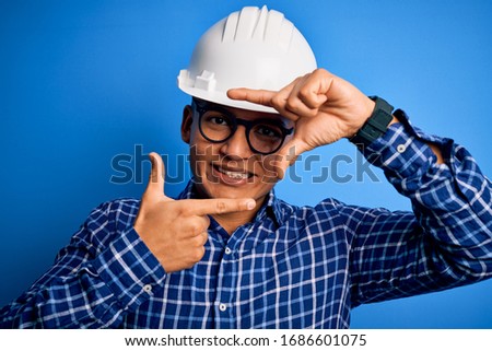 Young handsome engineer latin man wearing safety helmet over isolated blue background smiling making frame with hands and fingers with happy face. Creativity and photography concept.