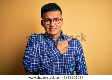 Young handsome latin man wearing casual shirt and glasses over yellow background Pointing aside worried and nervous with forefinger, concerned and surprised expression