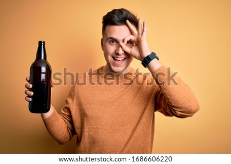Young handsome caucasian man drinking a bottle of beer over yellow background with happy face smiling doing ok sign with hand on eye looking through fingers