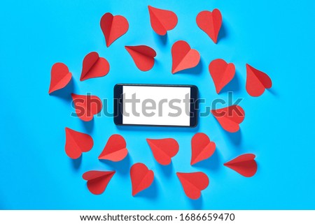 Black smartphone with isolated white screen for text, picture, photo and other graphics near scattered red paper hearts lies on blue table. Valentines day and love concept. Space for text. Top view