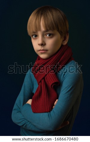 Portrait of a caucasian boy in a blue sweater and a borderous scarf on a dark blue background. Shot in 3/4. The child has long rustic hair, large light eyes. Look in the camera