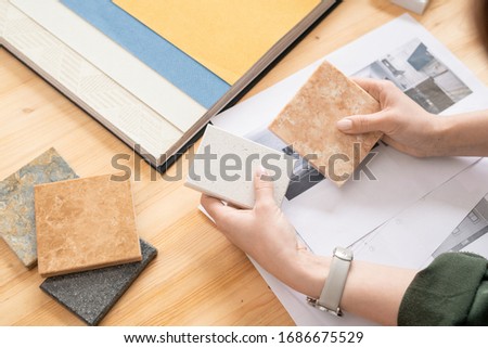 Hands of young female designer of interior comparing two samples of panels over papers with pictures of modern house or flat