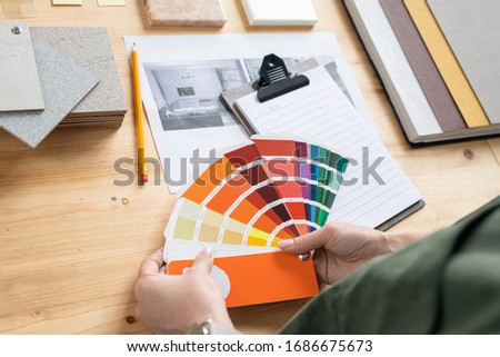 Hands of young female designer of interior holding color palette over desk while choosing suitable one for new order of client